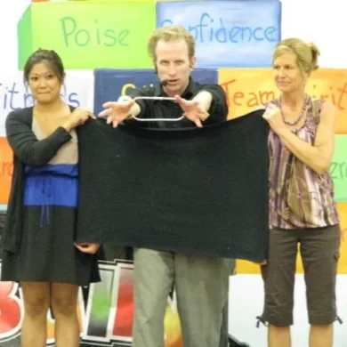 Performer doing a magic trick with a black cloth and two assistants during a school assembly