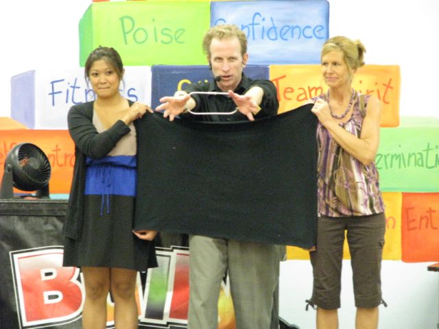 "School assembly performer demonstrates a magic trick with two adult volunteers in front of colorful character trait blocks."