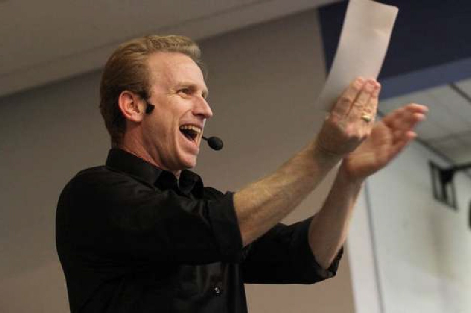 "School assembly presenter enthusiastically leading students with a big smile and a clapping gesture."