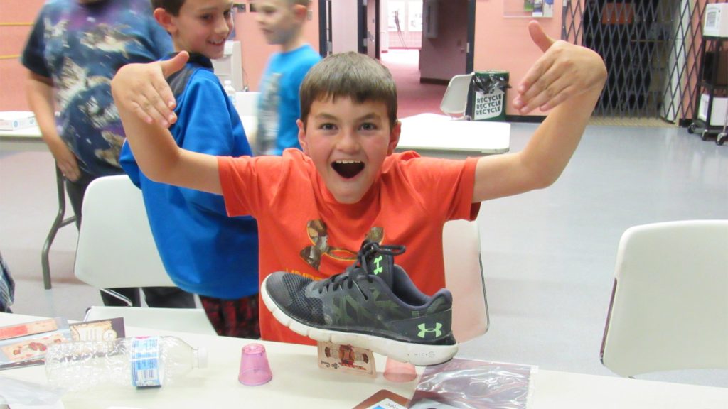 "Excited student proudly displays a shoe balanced on a bridge of paper cards during a hands-on school assembly activity."