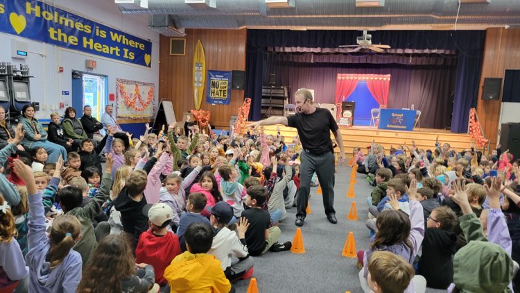 "School assembly presenter energizes a large group of students seated on the floor, all eagerly raising their hands in a decorated multipurpose room."